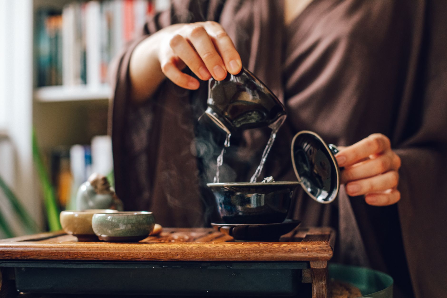 A small clay teapot pouring Chinese tea into thimble-sized tasting cups on a wooden tray