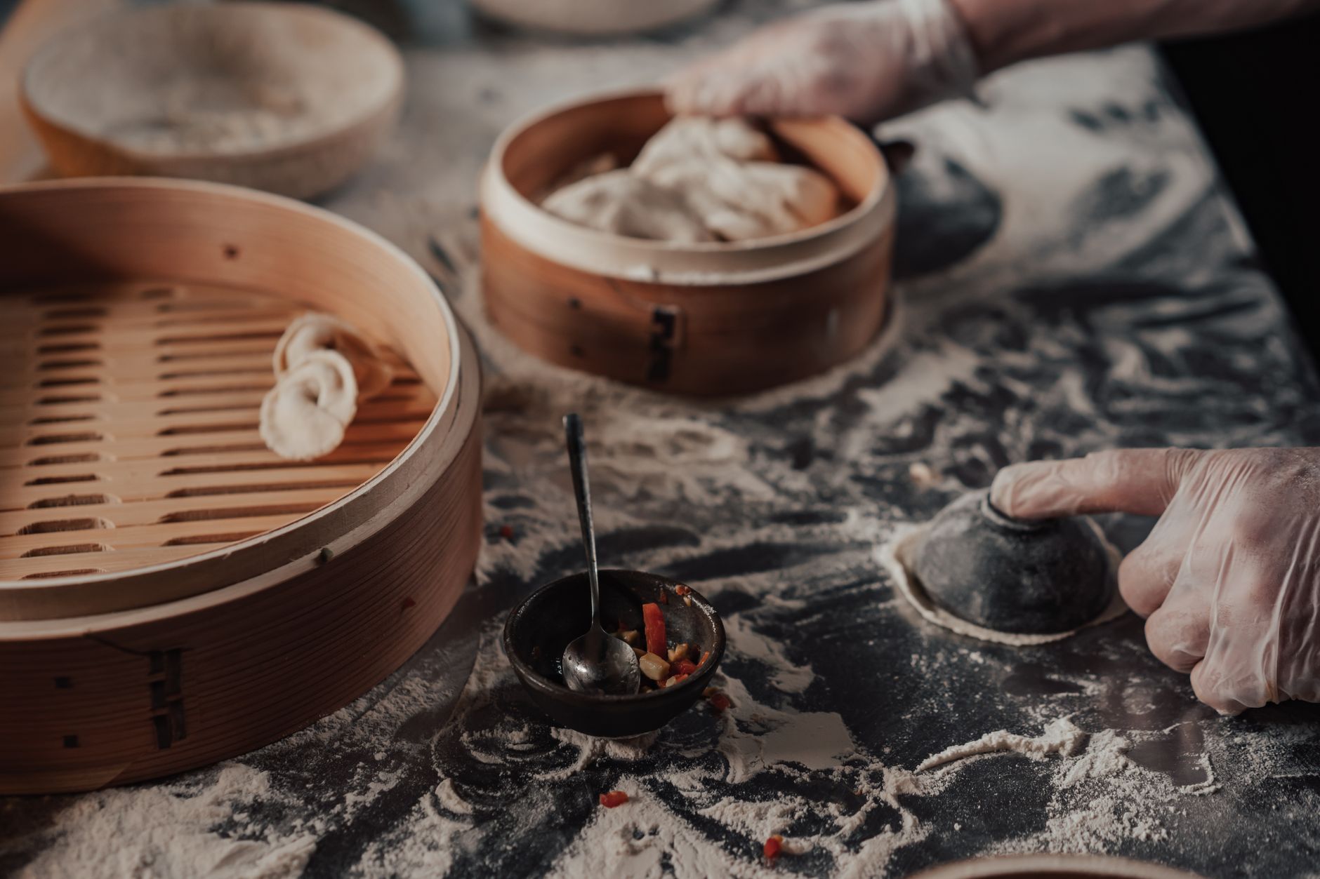Hands folding dumplings on a floured board beside stacked bamboo steamers