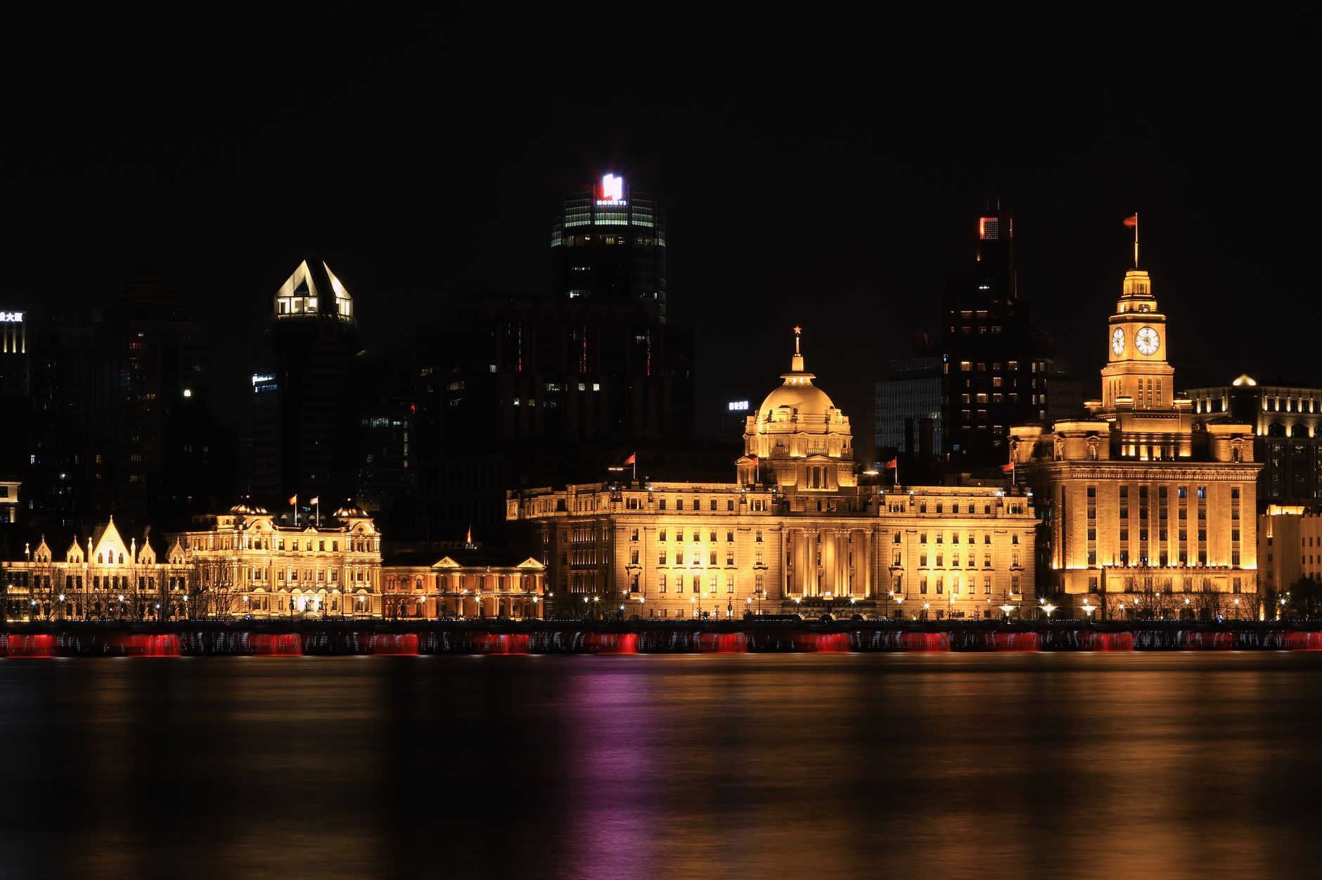 Illuminated 1920s stone buildings along the Bund seen across the Huangpu River at night