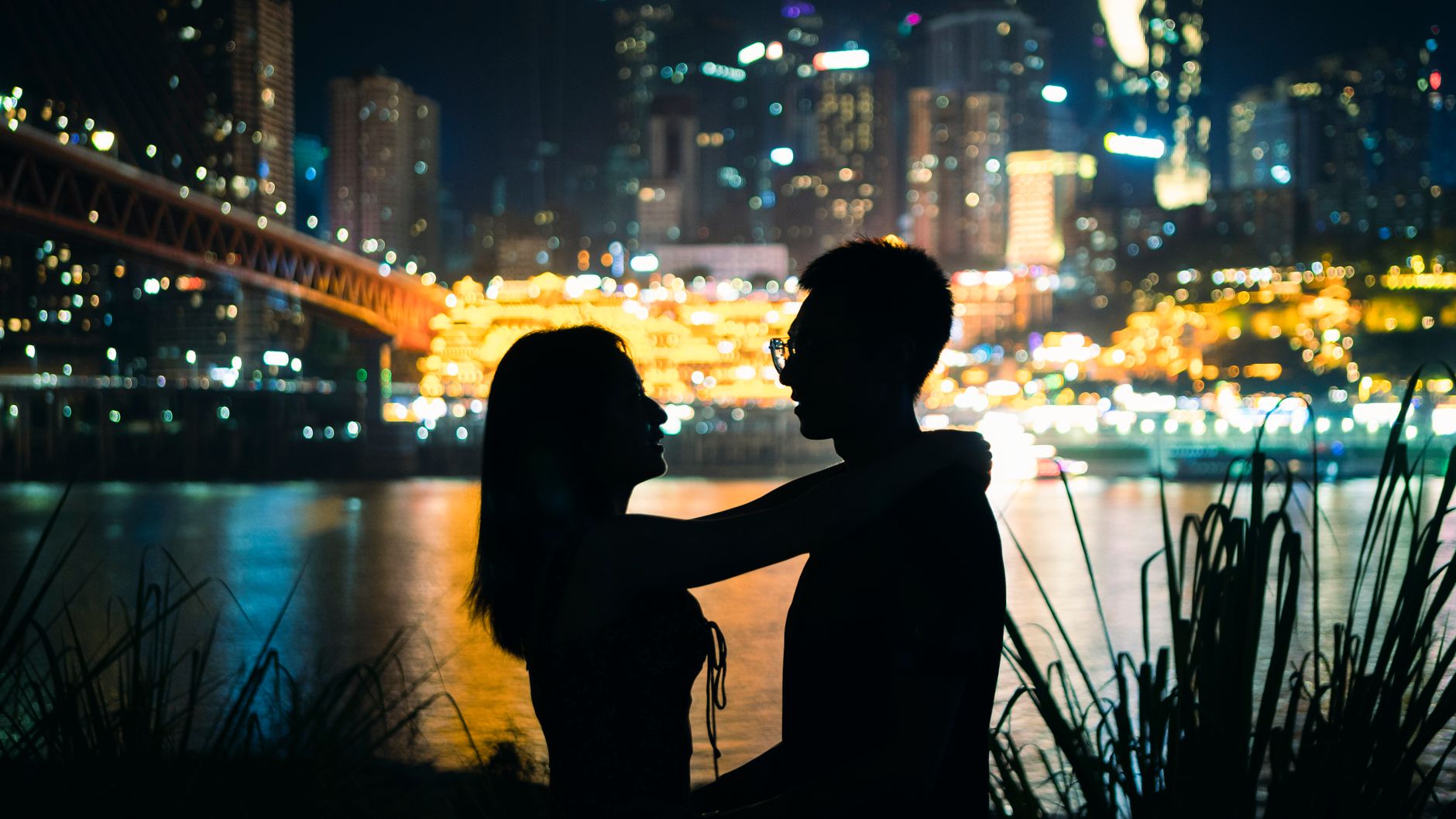 Silhouetted couple standing by a river with a lit city skyline behind them at night