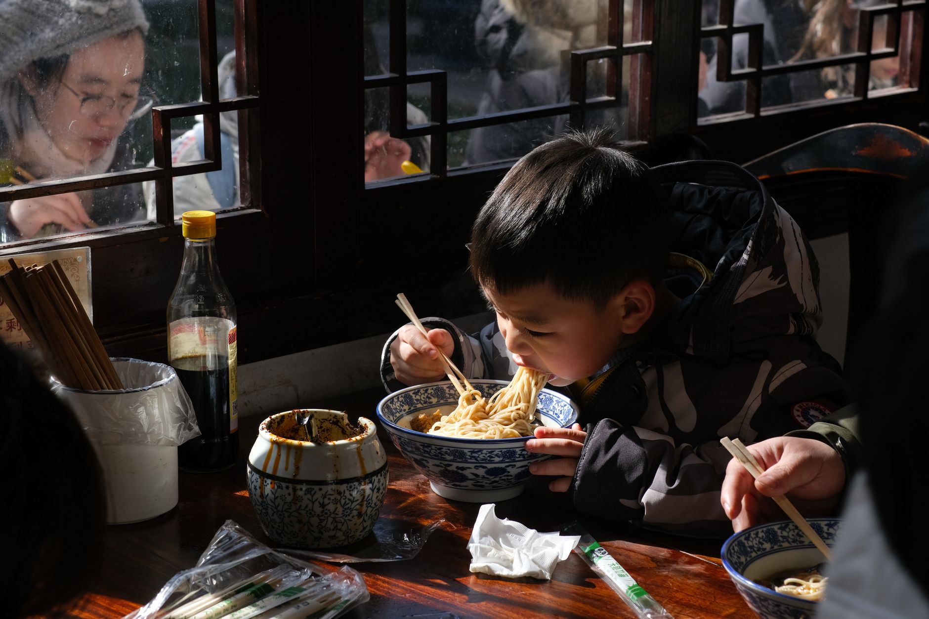 A young child eating a bowl of noodles with chopsticks at a simple restaurant table