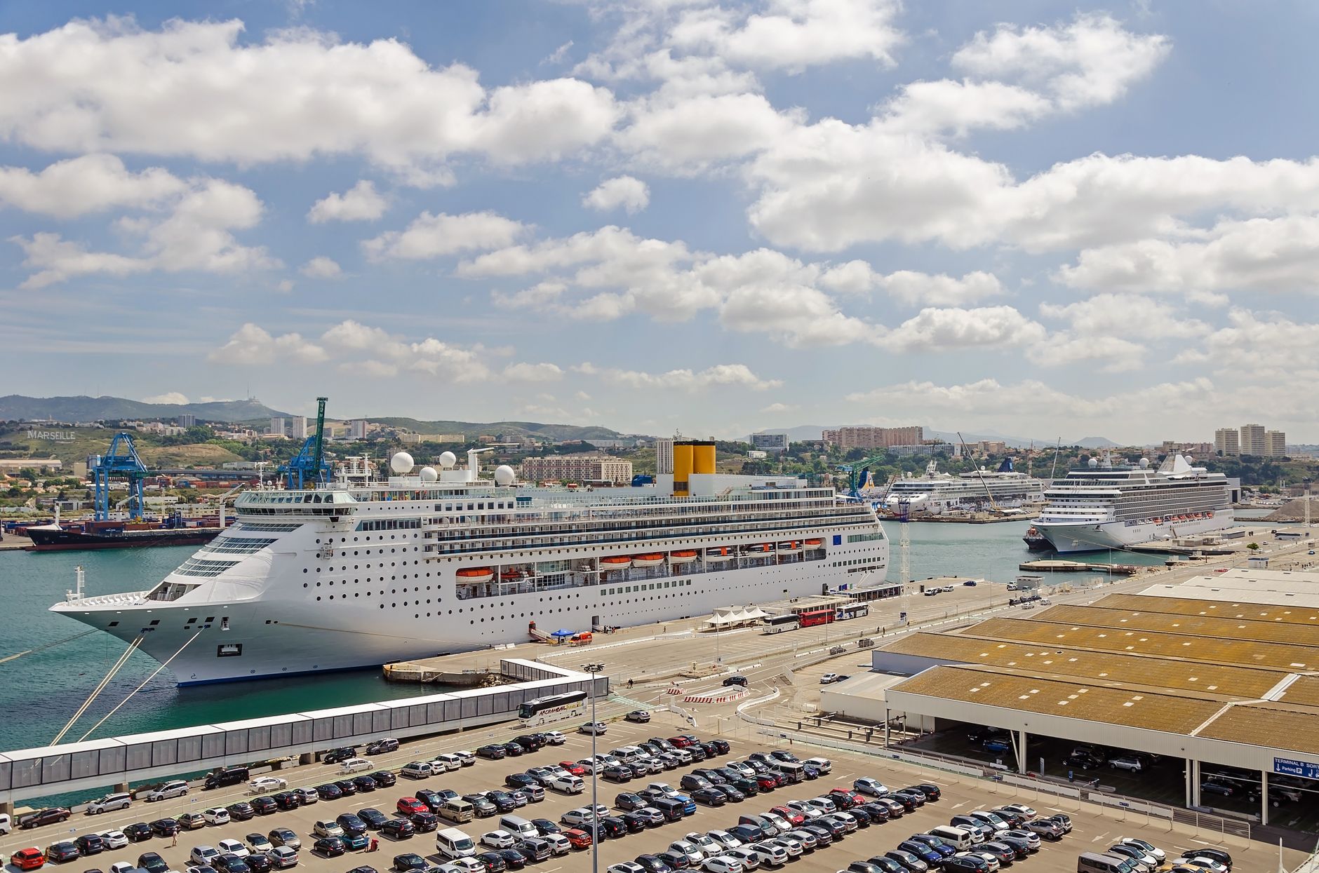 Cruise ships moored along a working port terminal with car parks and cranes behind