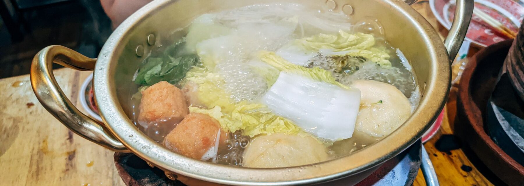 A brass hot pot of clear broth simmering over a burner with vegetables and fish balls