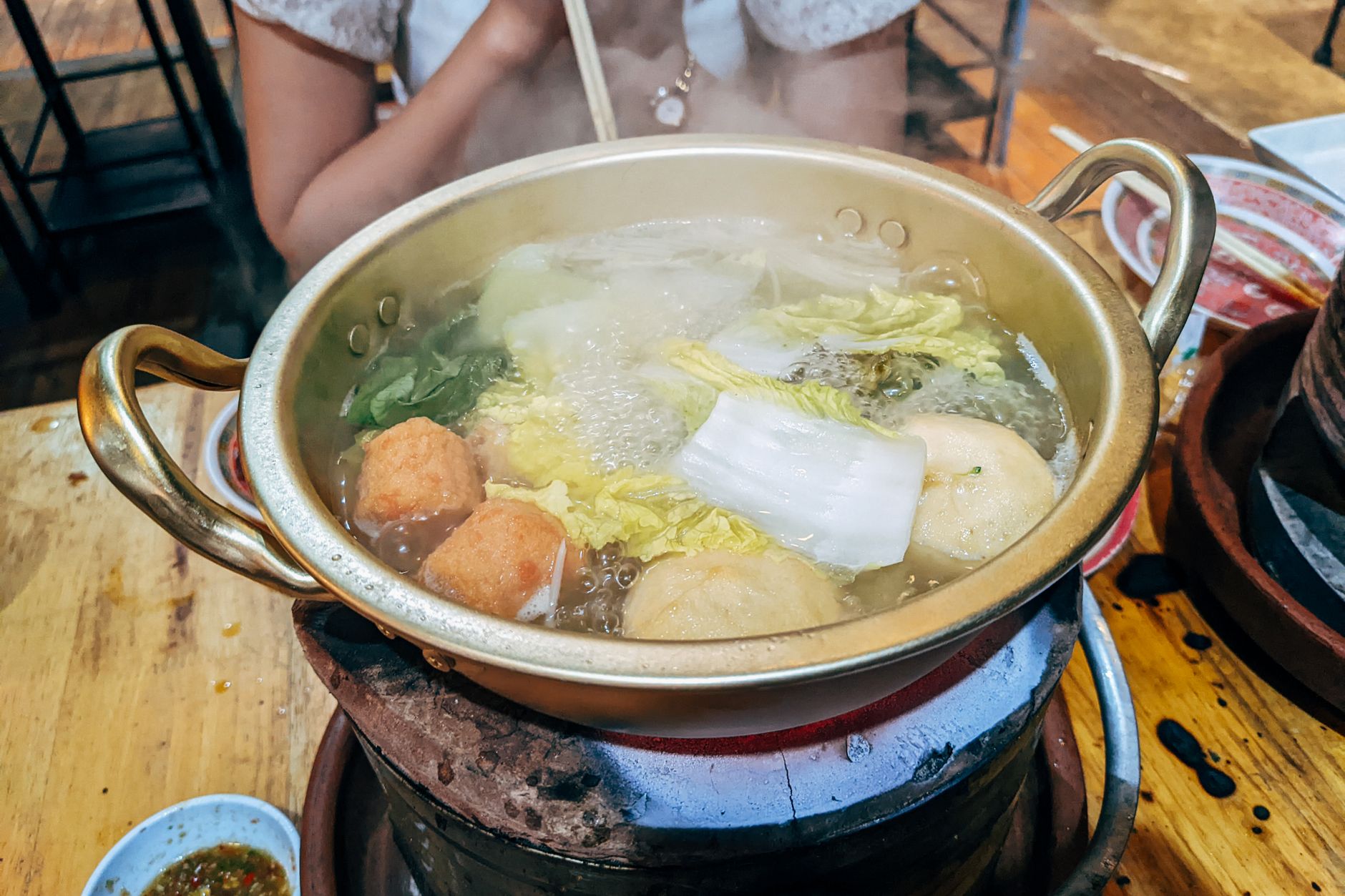 A brass hot pot of clear broth simmering over a burner with vegetables and fish balls