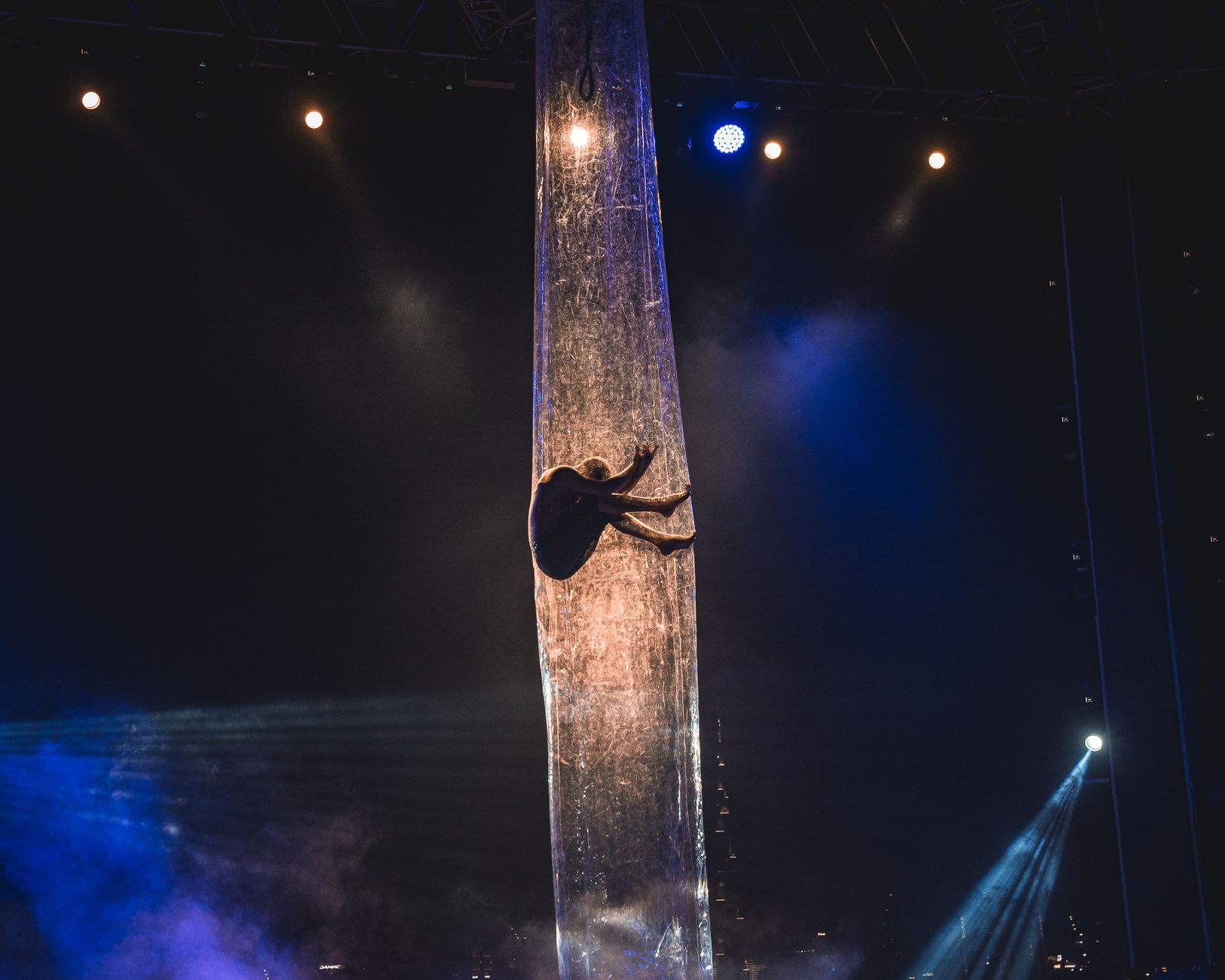 A single aerial performer climbing a fabric silk high above a stage in coloured light