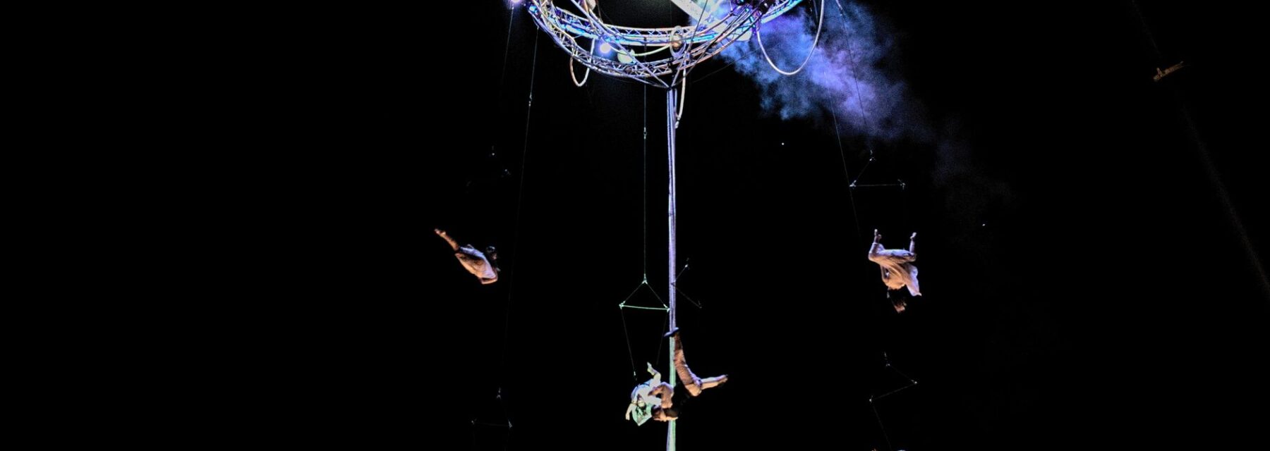 Aerial acrobats suspended from a lit rig above a darkened stage