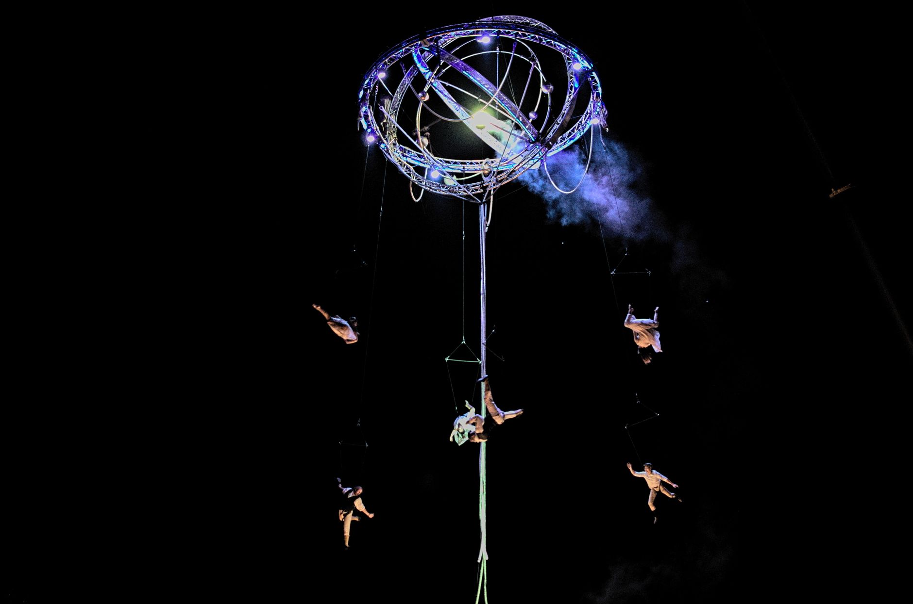 Aerial acrobats suspended from a lit rig above a darkened stage
