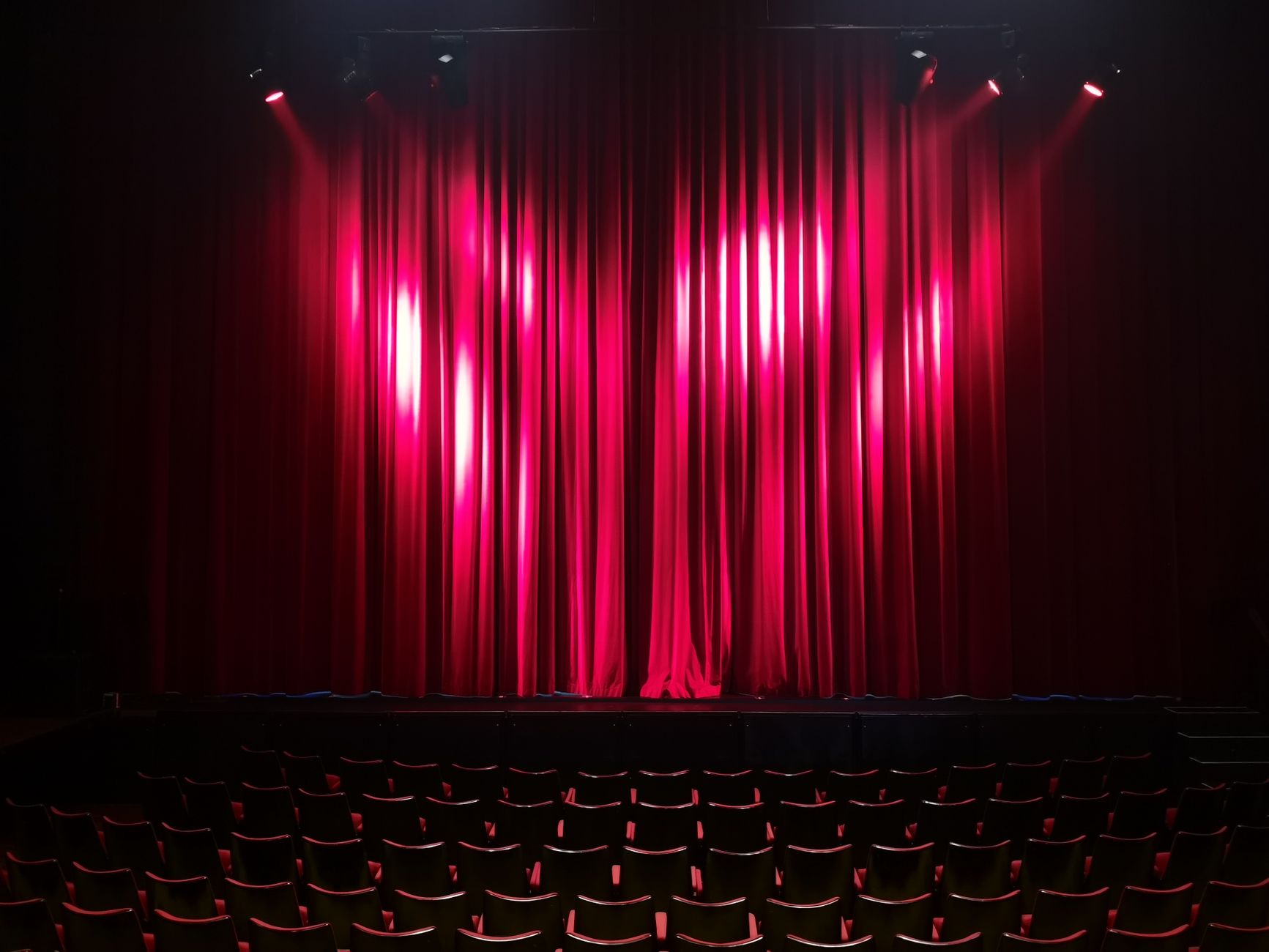 An empty theatre stage behind a lit red curtain with rows of seats in front