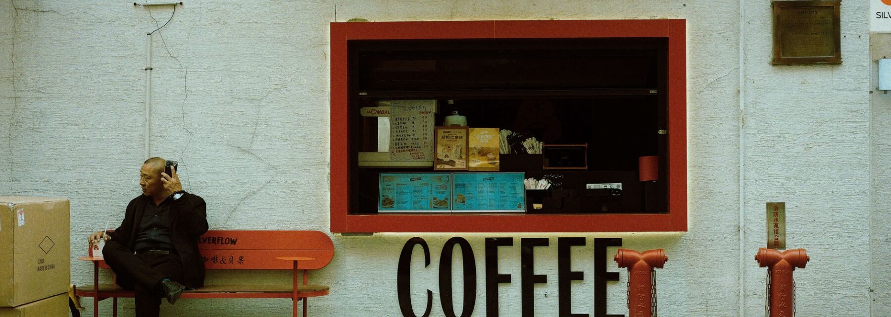 A man sitting on a bench outside a small street-front coffee shop in Shanghai with a hatch window