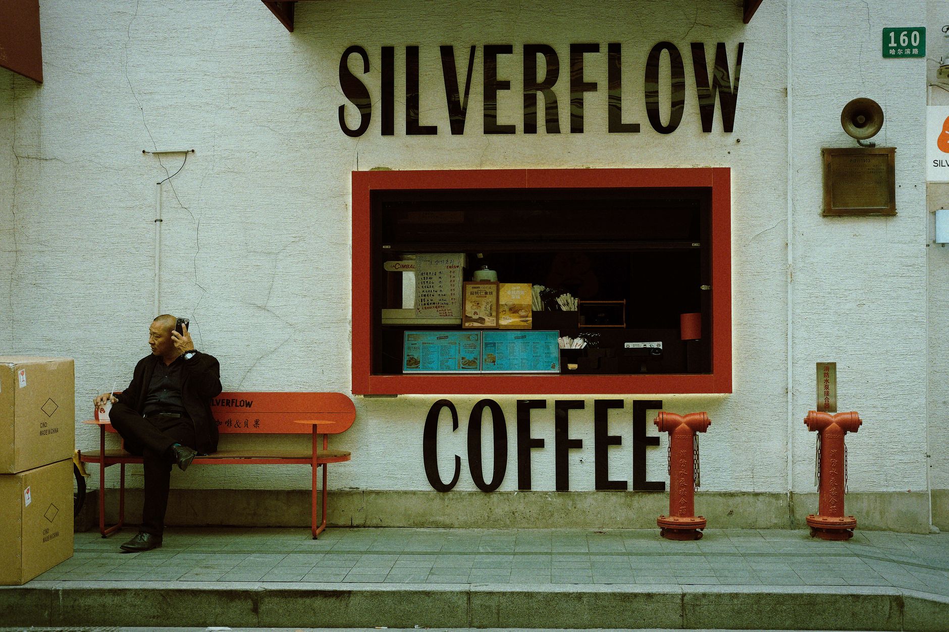 A man sitting on a bench outside a small street-front coffee shop in Shanghai with a hatch window