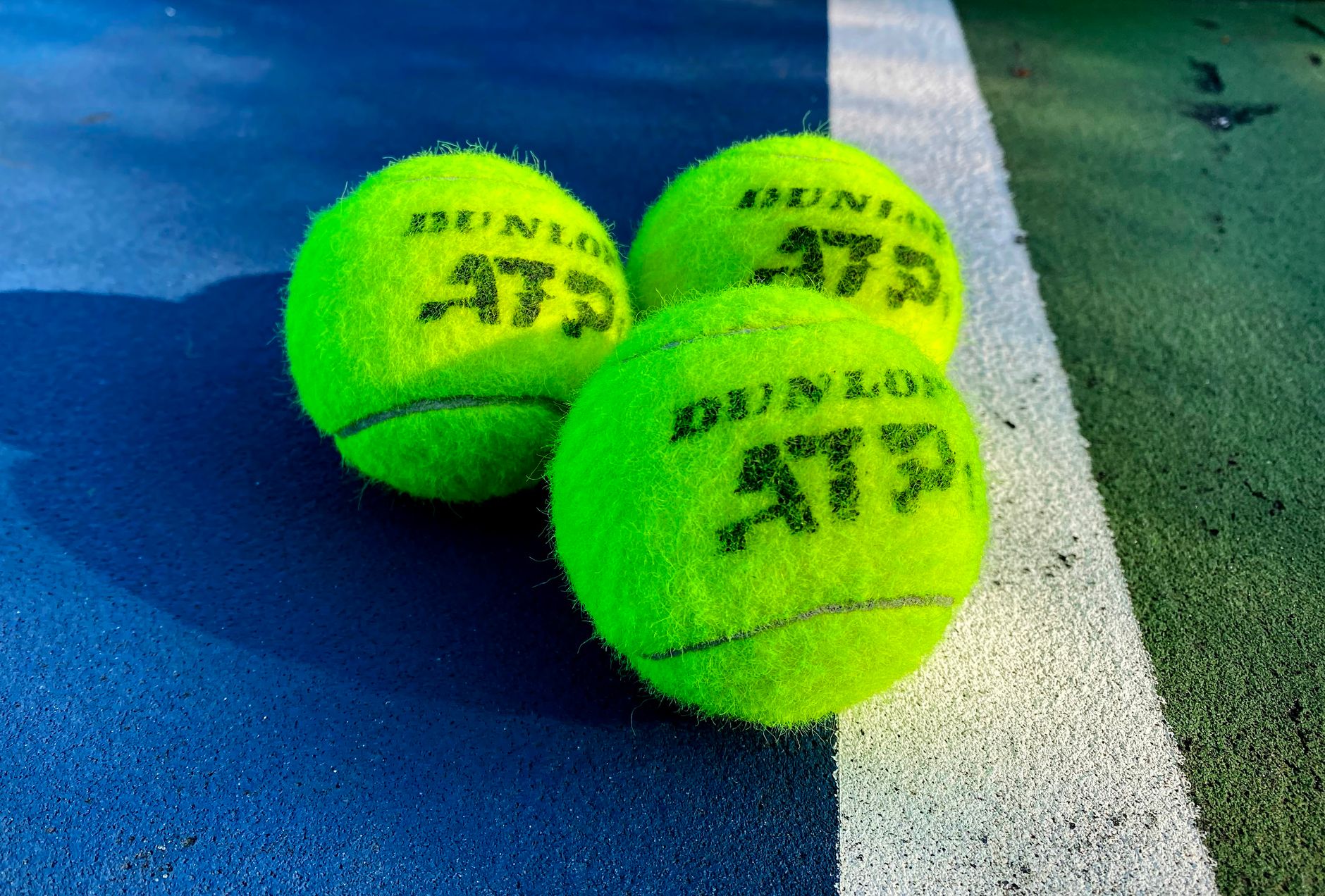 Three ATP-branded tennis balls on a blue hard court beside a white line