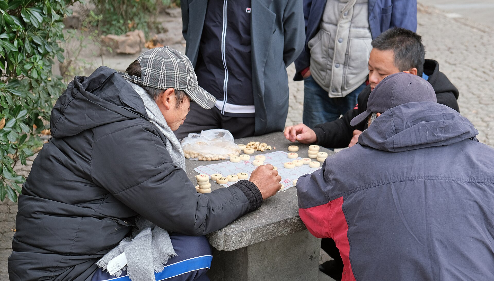 Older men in winter coats gathered around a board game on a stone table in People's Park