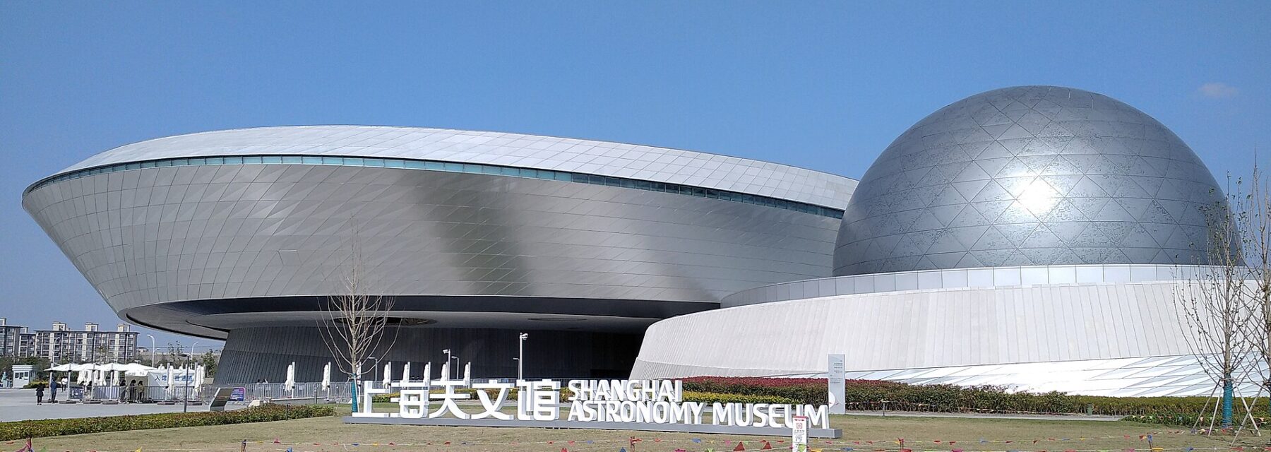 The curved white building of the Shanghai Astronomy Museum with its silver sphere on the roof, behind a lawn and a sign reading Shanghai Astronomy Museum