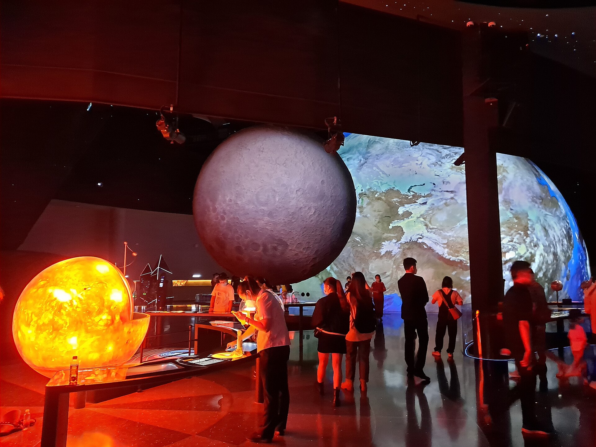 Visitors in a dark hall with a large glowing Sun model, a grey Moon model and a projected Earth