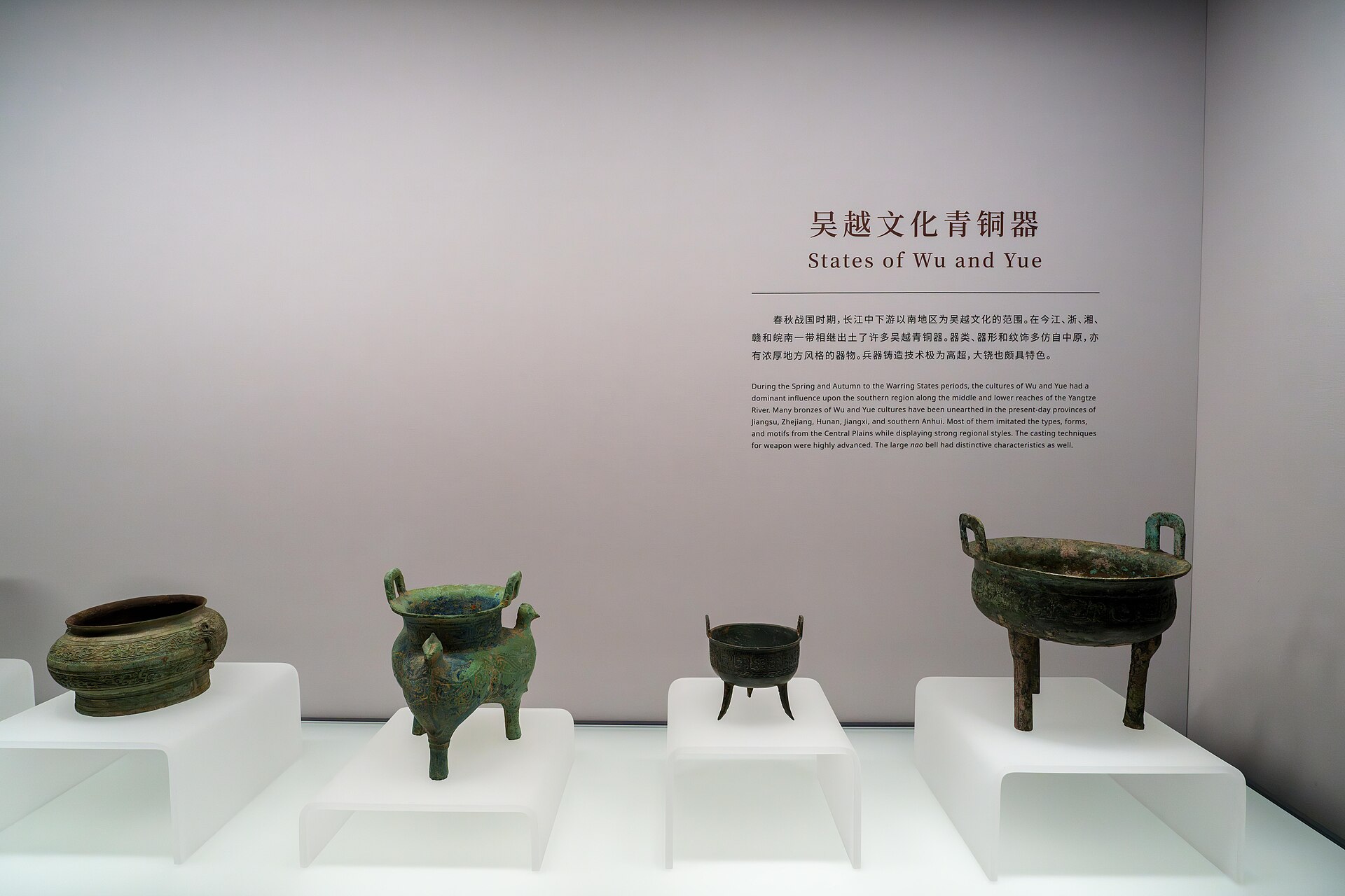 Four ancient bronze vessels with legs and handles displayed on white plinths beneath a wall panel headed States of Wu and Yue