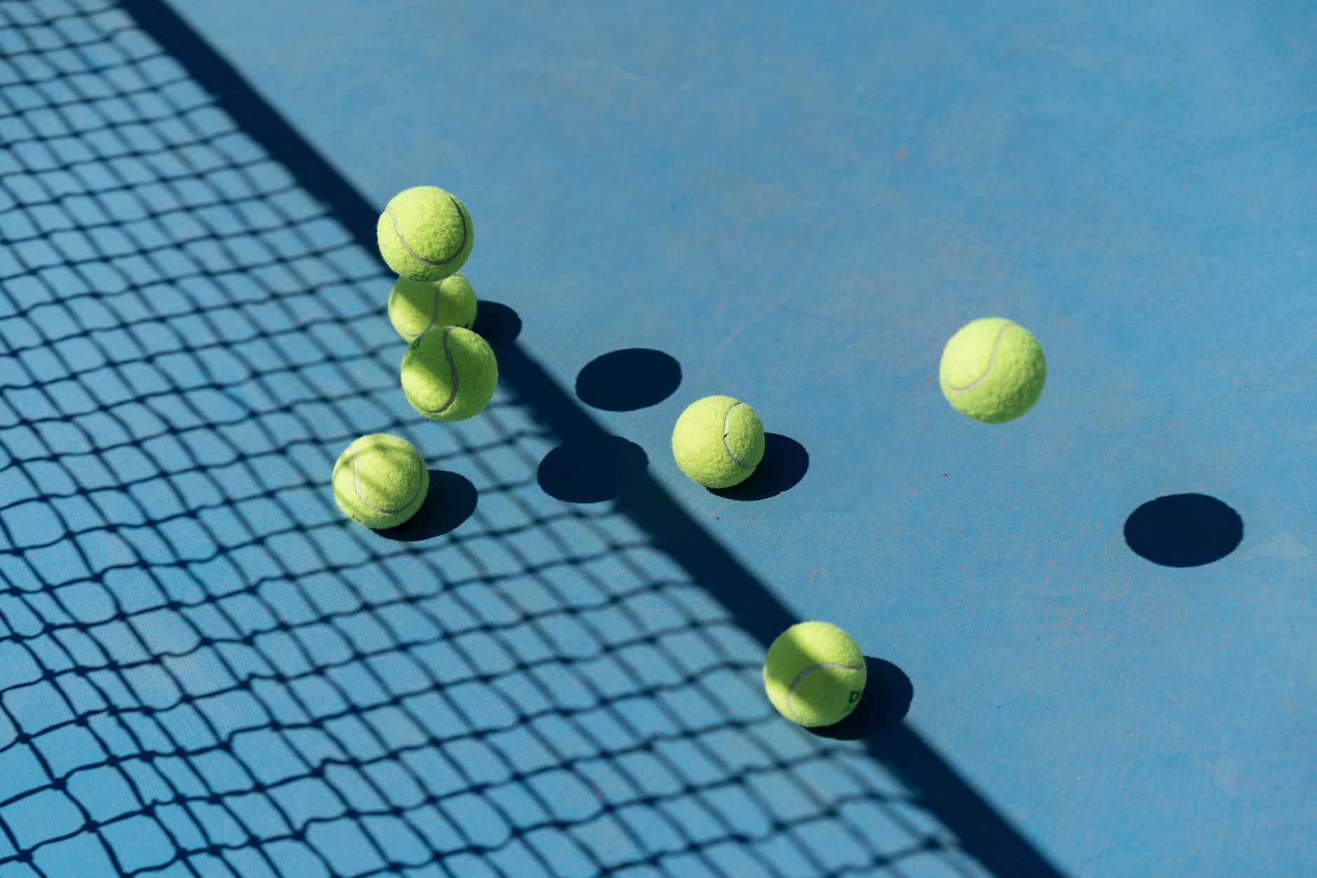Tennis balls scattered across a blue hard court with the shadow of the net falling over them