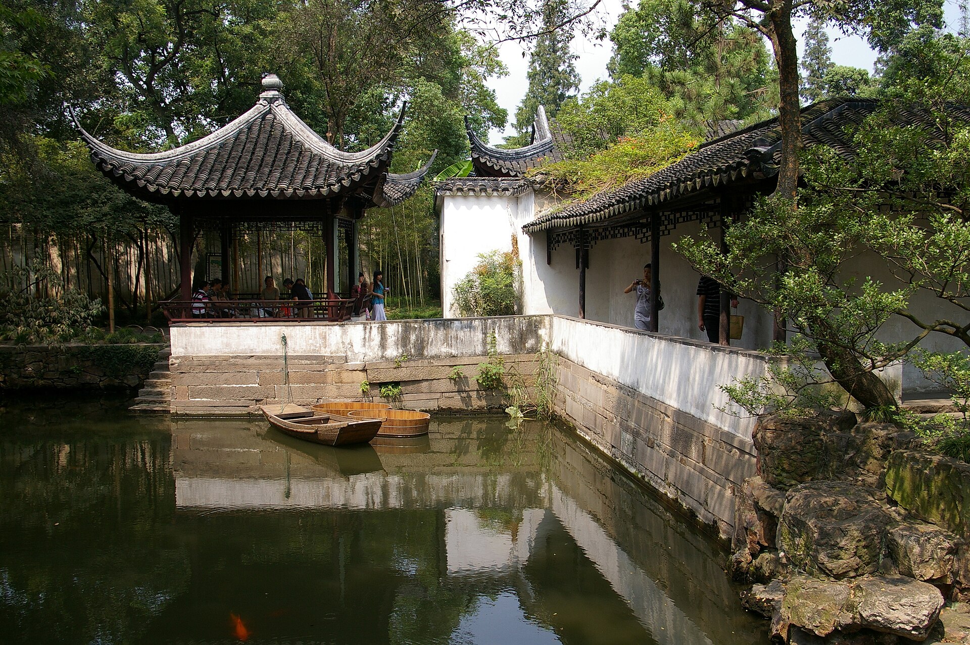 A tiled pavilion reflects in a Suzhou garden pond beside white corridor walls and two small wooden boats.